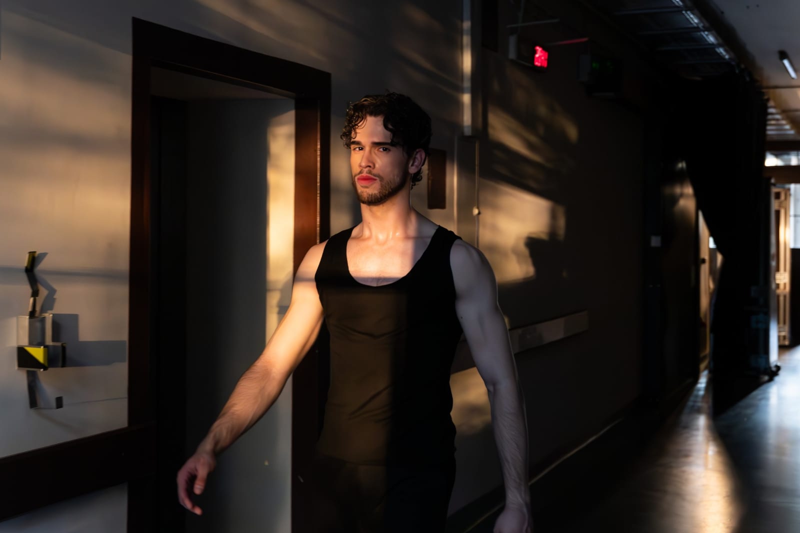 A dancer in a black tank top walking through a theatre corridor in warm golden light