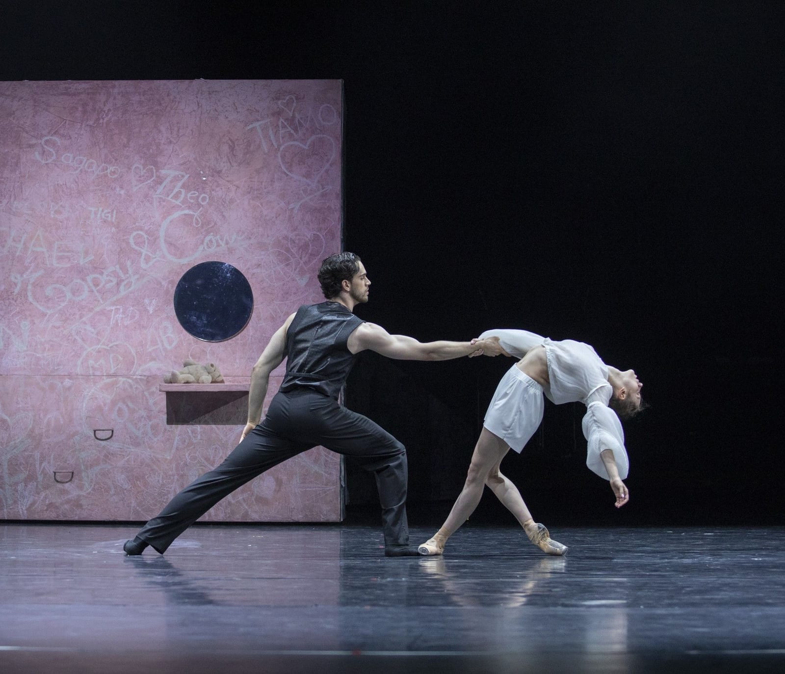 Scene from R+J by Robert Bondara: a dancer as Paris lunges and holds a female dancer's hand as she arches back on pointe, on a pink graffiti-covered set