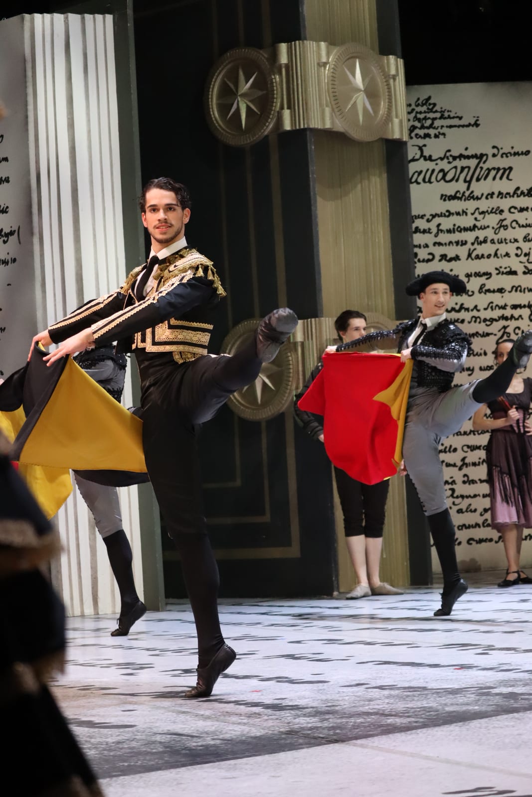 Scene from Don Quixote: Espada in a black and gold toreador jacket kicks one leg high while holding a yellow and black cape, with other toreadors behind him