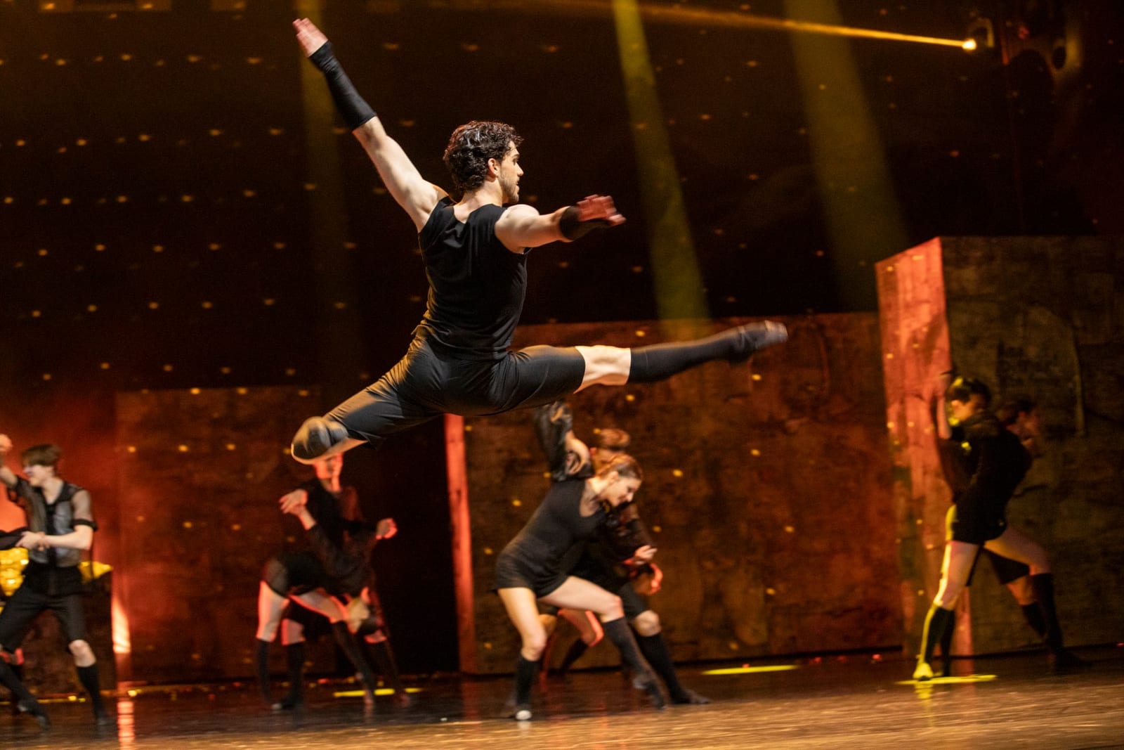 Scene from R+J by Robert Bondara: a dancer as Tybalt caught mid-leap on a gold-lit stage, with ensemble dancers behind him
