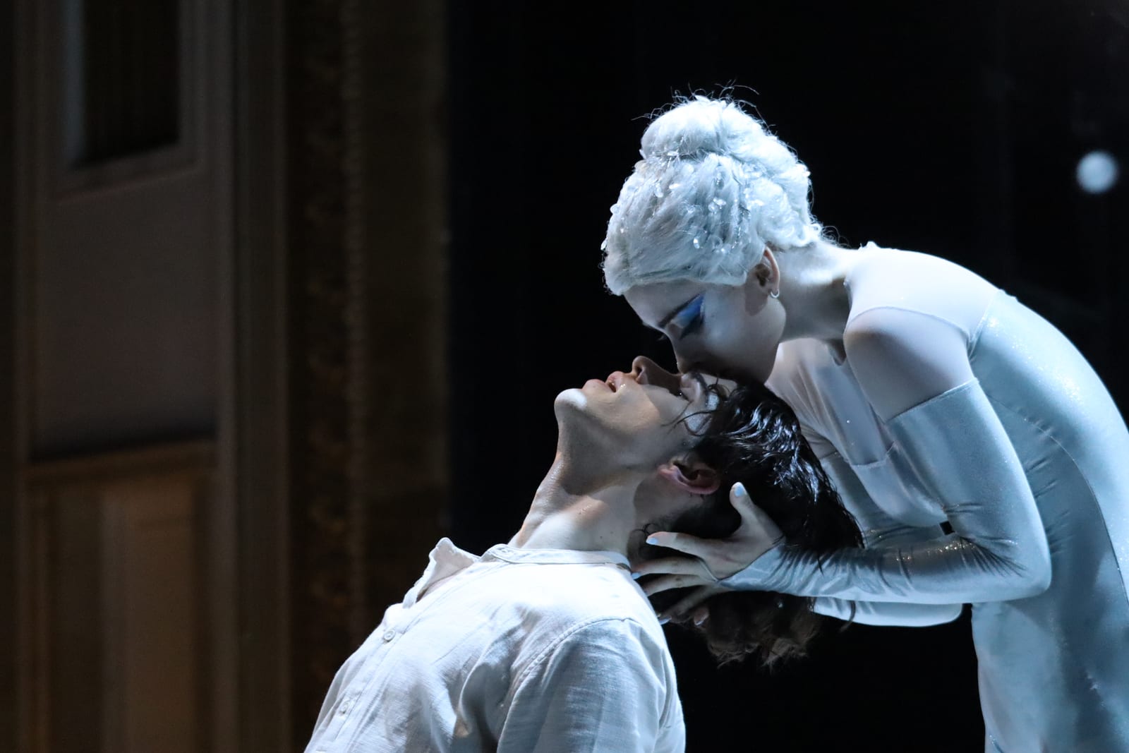 Scene from Snow Queen by Robert Bondara: a dancer in a white wig and silver-white costume bends to kiss the forehead of Kaj, who leans back in a white shirt