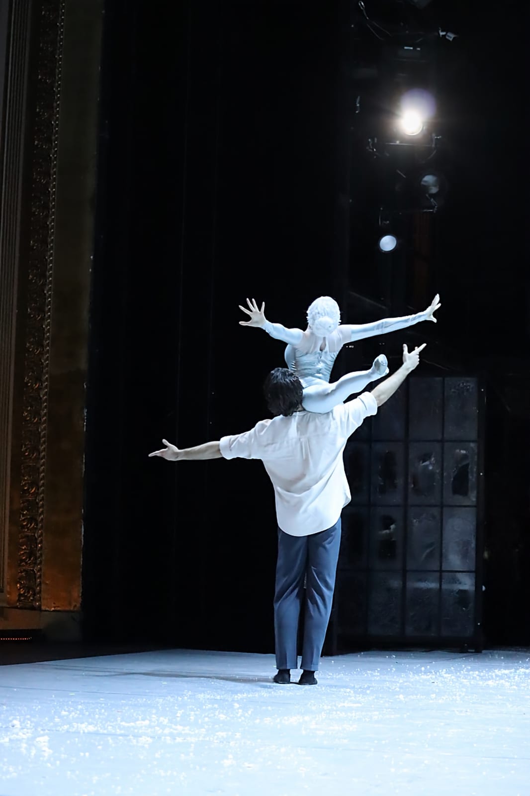 Scene from Snow Queen by Robert Bondara: Kaj, in a white shirt seen from behind, lifts a dancer in a silver-white costume onto his shoulder on a snow-covered stage